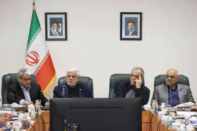 Four men in suits sit at a conference table with microphones, with an Iranian flag and two framed portraits hanging on the wall behind them.