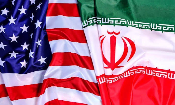 A gray-haired, bearded man wearing glasses, a light blue shirt, and a dark blue suit stands before a partially visible Iranian flag.