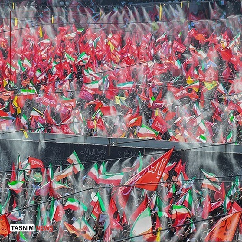 Tasnim News logo visible. A large crowd waves red, green, and white flags through haze and power lines at an outdoor gathering.