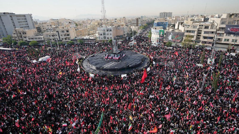 A massive crowd carrying red flags surrounds a central monument in an urban square, with multi-story buildings visible in the hazy background.