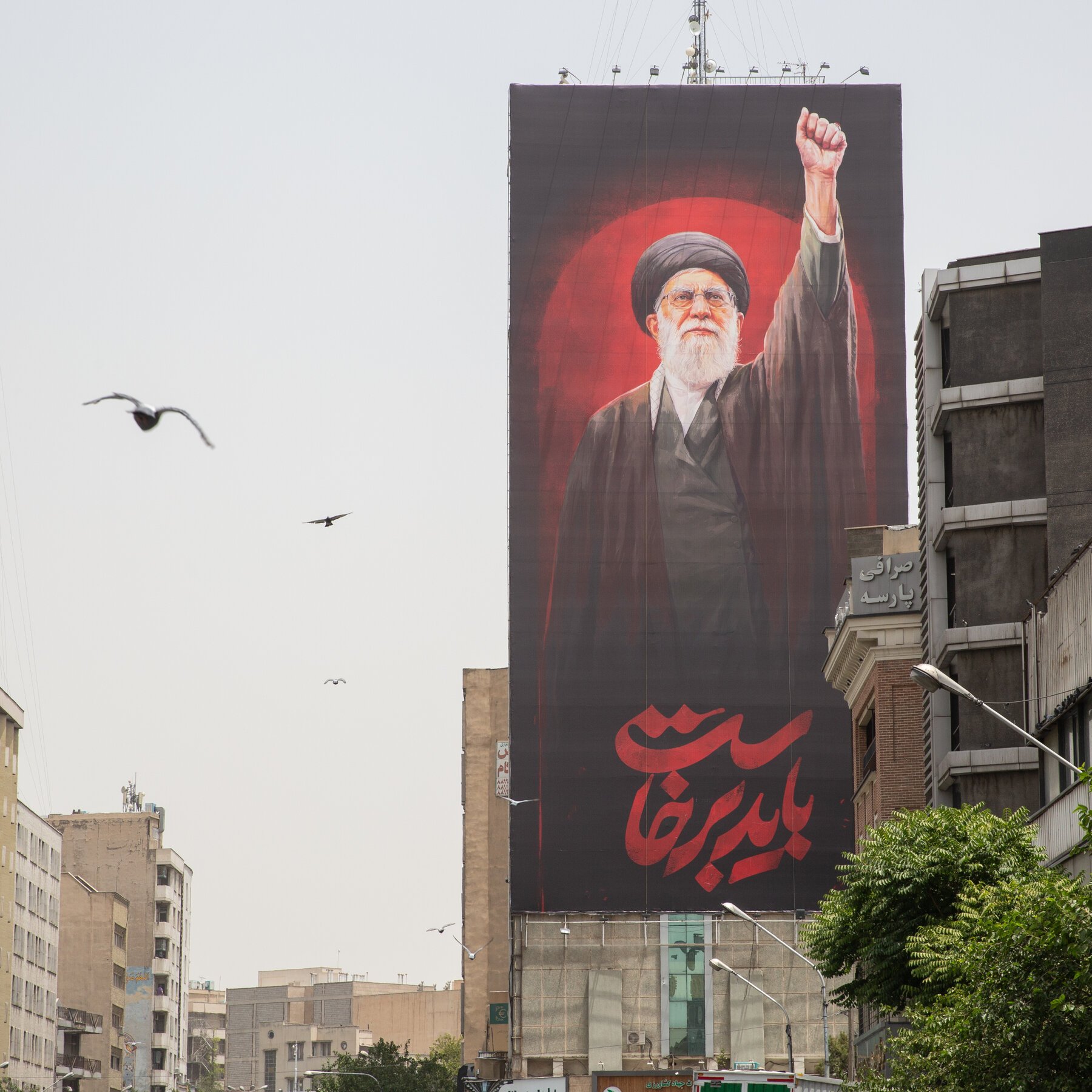 Aerial view of a massive crowd filling an urban street, waving red and Iranian flags between buildings, with Farsi text overlay in the corner.