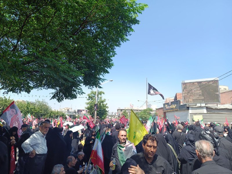 A crowd gathers on a tree-lined street, waving Iranian flags and holding a portrait placard beneath a clear blue sky.