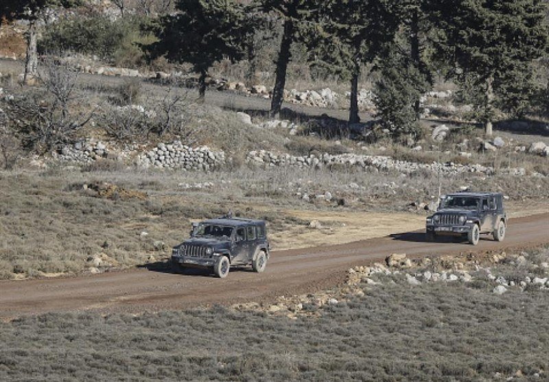 Two military-style vehicles travel along a dirt road through a rural landscape with stone walls, sparse trees, and rocky terrain.