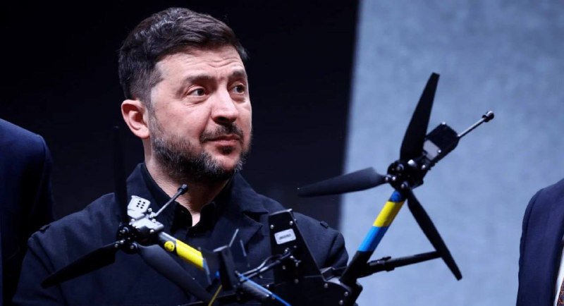 A dark-haired, bearded man in a black shirt stands behind a display of two drones with yellow and blue components.