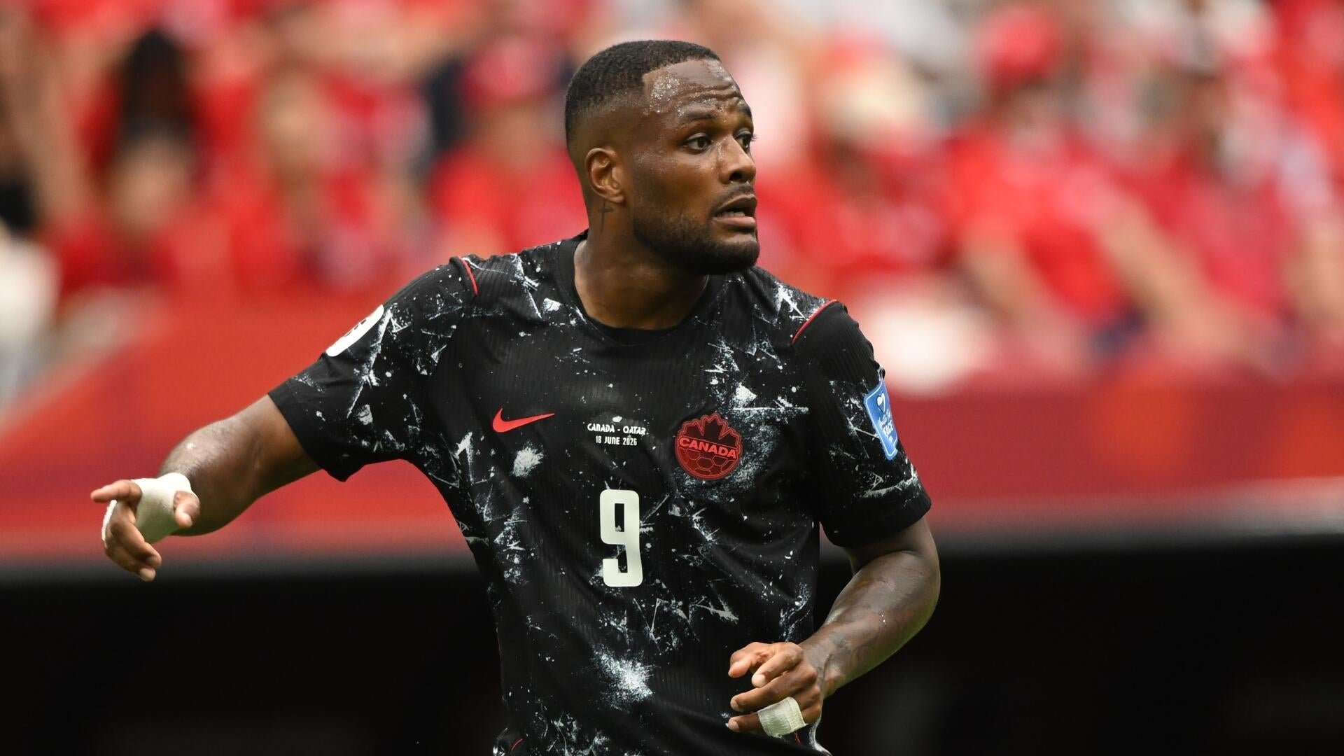 A soccer player wearing a red-and-white striped jersey with the number 20 and a USA crest stands in a stadium with a blurred crowd behind him.