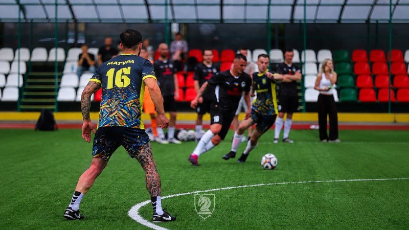 A soccer player wearing jersey number 16 with "МАРУЩАК" kicks a ball on a green field while opponents and spectators watch from stadium stands.