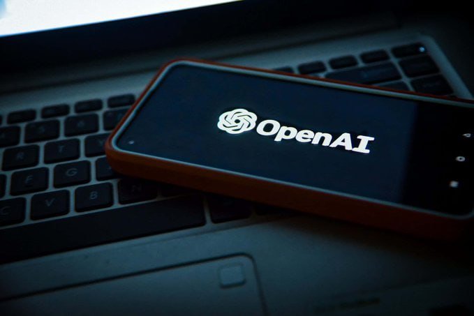A smartphone with a red case displaying the OpenAI logo on its screen rests atop a laptop keyboard.