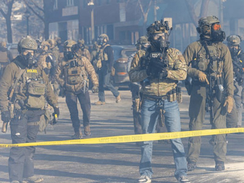 Heavily armed individuals in tactical gear, gas masks, and helmets walk along a street behind yellow "DO NOT CROSS" tape.