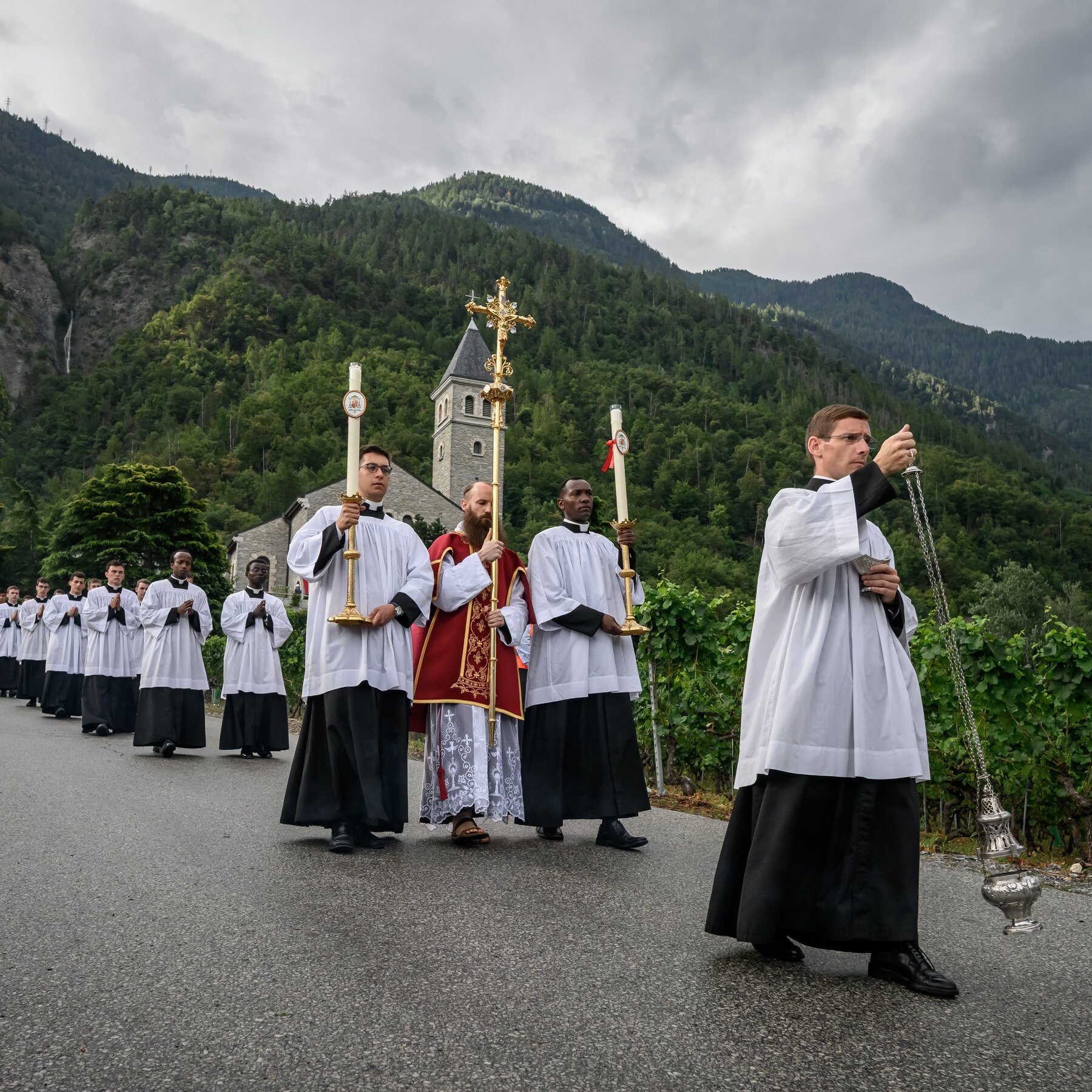 Clergy in white and black vestments process along a road, carrying a golden cross, candles, and a thurible, with a stone church and forested mountains in the background.