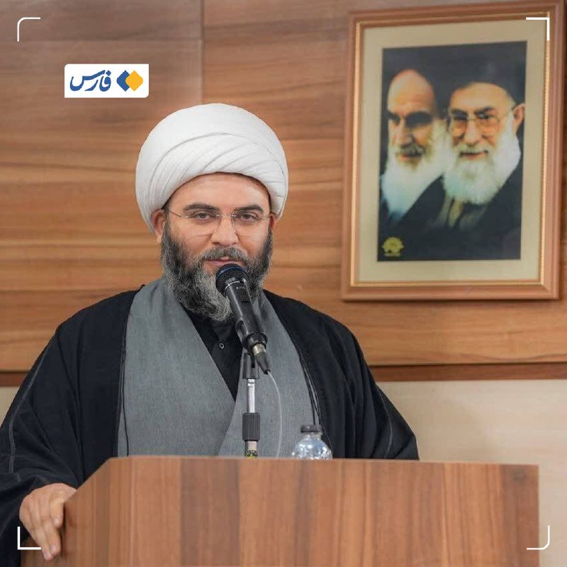 A bearded man in clerical attire and white turban speaks at a microphone behind a wooden podium, with a framed portrait of two bearded men hanging on the wood-paneled wall behind him.
