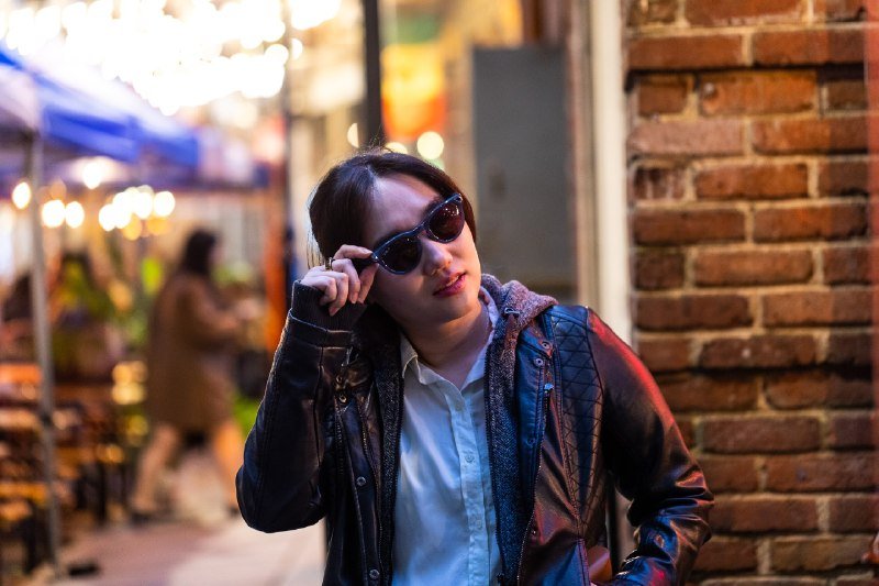 A woman in sunglasses and a black leather jacket stands against a brick wall at night, with blurred lights and a passing pedestrian in the background.