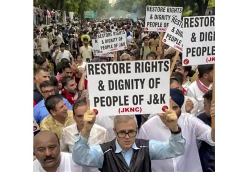 A large crowd of protesters marches outdoors, holding multiple signs reading "RESTORE RIGHTS & DIGNITY OF PEOPLE OF J&K (JKNC)."