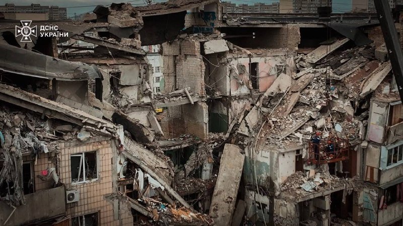 A multi-story residential building lies in ruins, with collapsed floors, exposed interiors, and scattered debris, bearing a logo reading "ДСНС України" in the upper left.