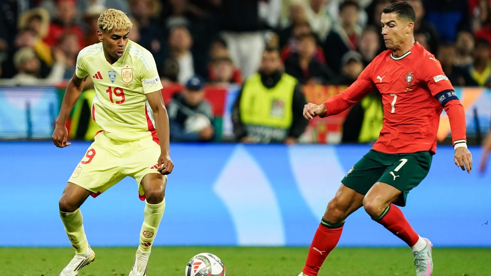 Two soccer players, one in a yellow kit (#19) and another in a red kit (#7), compete for a Nations League match ball on a pitch before a stadium crowd.