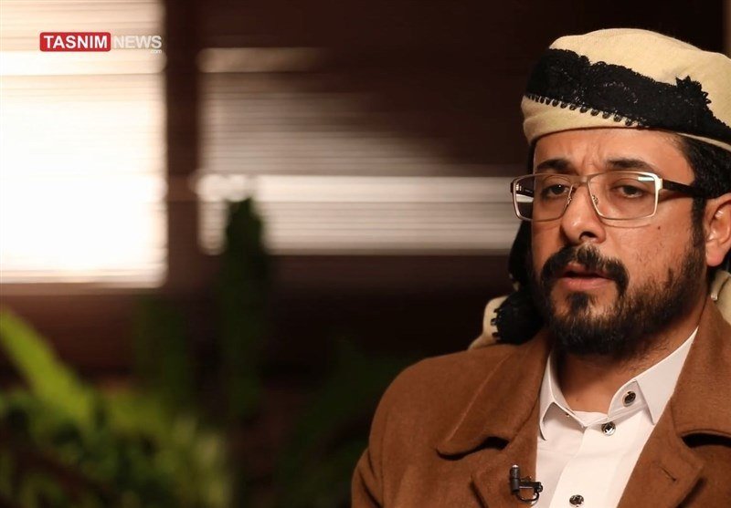 A bearded man wearing glasses, a beige and black head covering, and a tan jacket speaks indoors, with window blinds and a plant visible in the background.