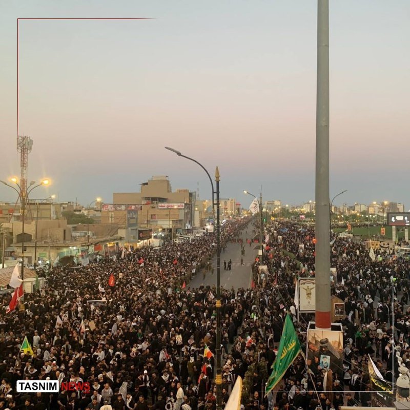 A large crowd fills a wide city street at dusk, with people carrying various flags and banners while buildings line the route.