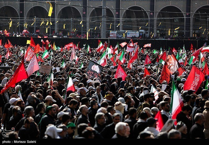 A massive crowd waves red flags and Iranian flags at a large outdoor rally, with a portrait banner visible in the distance.