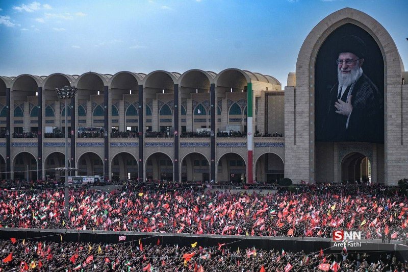 Women in black chadors hold portraits of bearded men in turbans and red flags with Persian script during an outdoor gathering under a blue sky.