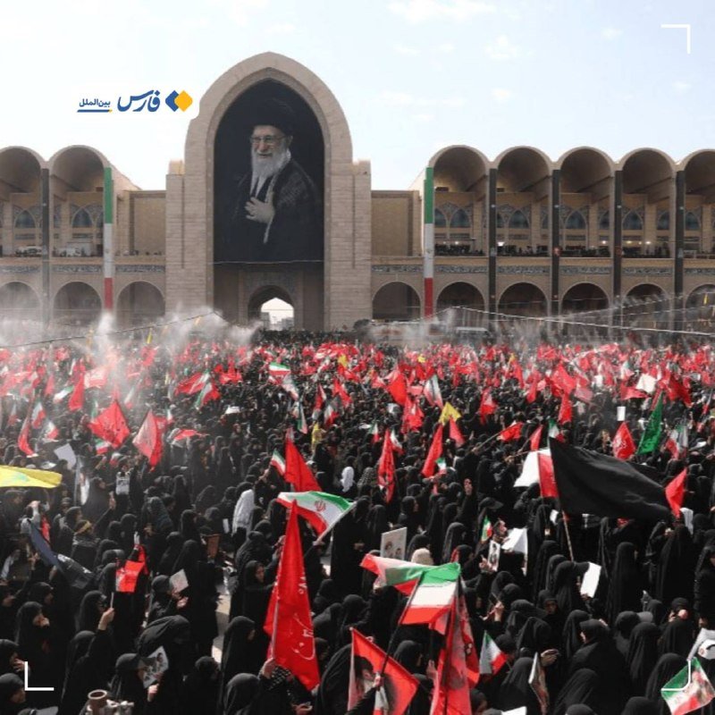 Four men sit at a conference table with microphones, papers, and bottles, beneath an Iranian flag and two framed portraits on the wall.