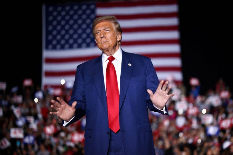 A man in a blue suit and red tie stands with arms outstretched before a large American flag backdrop and a crowd holding campaign signs at a nighttime rally.