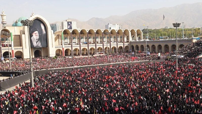An elderly man with white hair and glasses, wearing a dark suit and blue patterned tie, gestures with his hands while speaking in a formal room.