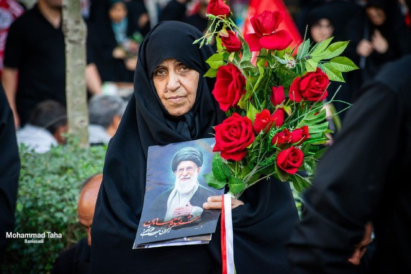 A woman in a black hijab holds a bouquet of red roses and a portrait of a bearded man in clerical attire, surrounded by people in black clothing.