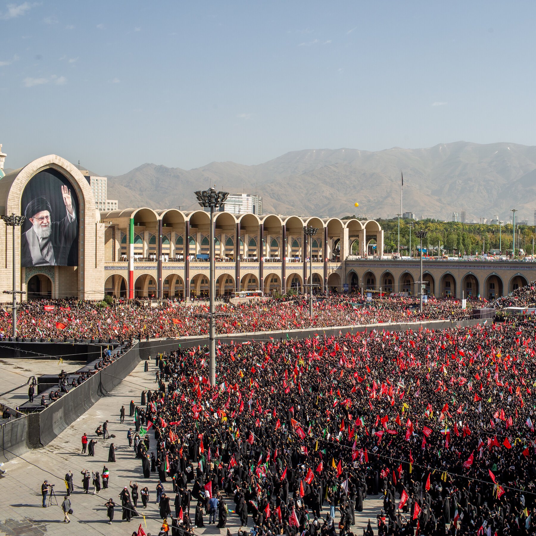 A crowd of women in black chadors hold portraits, red flags with Arabic script, and signs during an outdoor daytime demonstration under a blue sky.