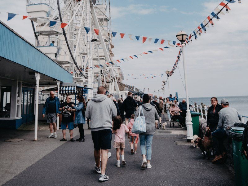 A crowd of people strolls along a seaside promenade beside a large Ferris wheel, with red, white, and blue bunting strung overhead and the ocean visible in the background.