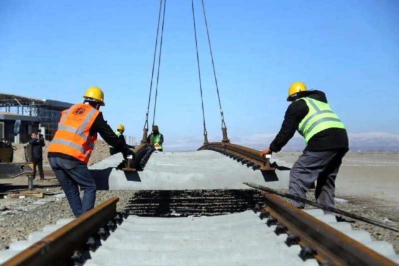 Workers in hard hats and high-visibility vests guide a suspended concrete panel over railway tracks during construction.