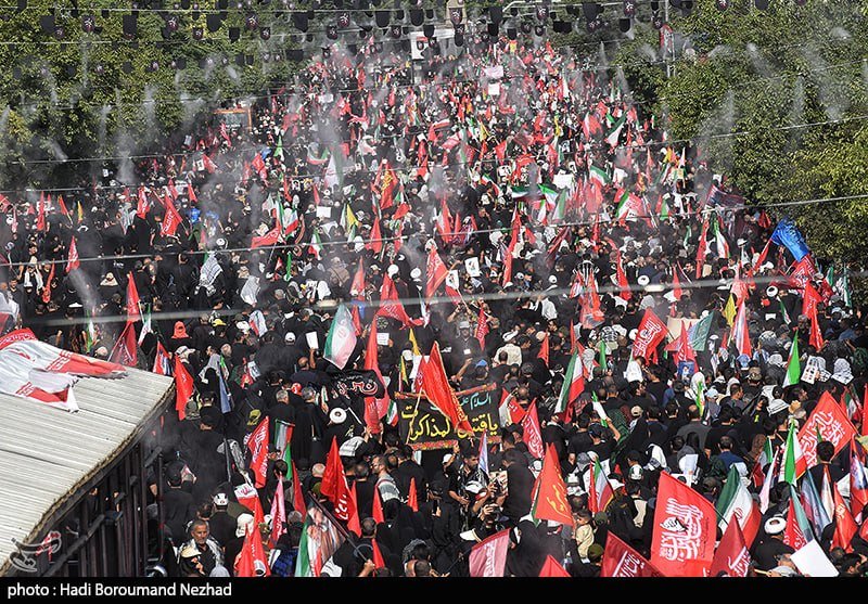 A large crowd marches down a tree-lined street waving numerous red, black, white, and green flags and banners bearing Arabic script.