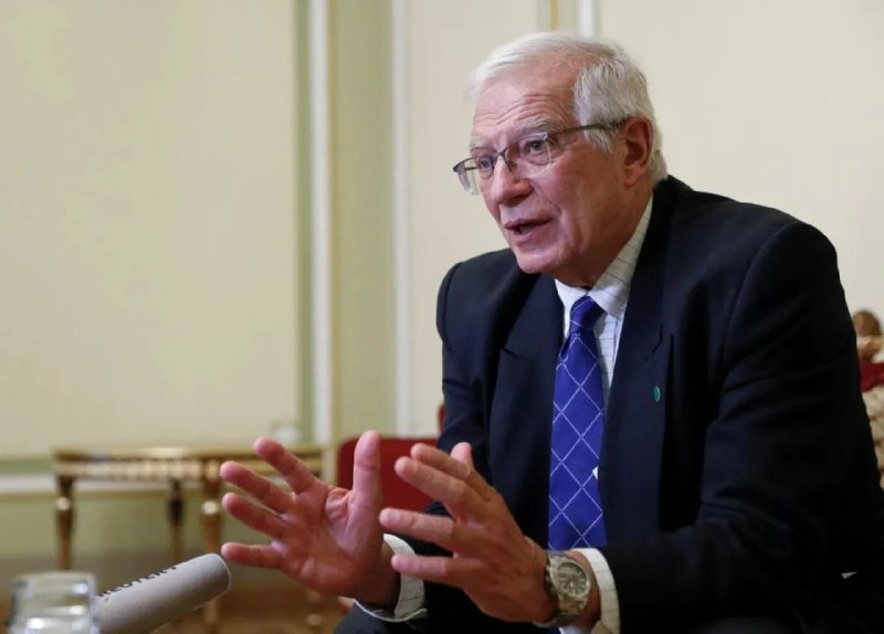 An older man with white hair and glasses, wearing a dark suit and blue patterned tie, speaks while gesturing with both hands in a formal indoor setting.