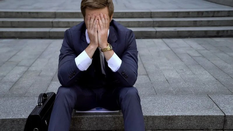 A man in a dark suit sits on stone steps with his face buried in both hands, a black briefcase beside him and a folder on his lap.