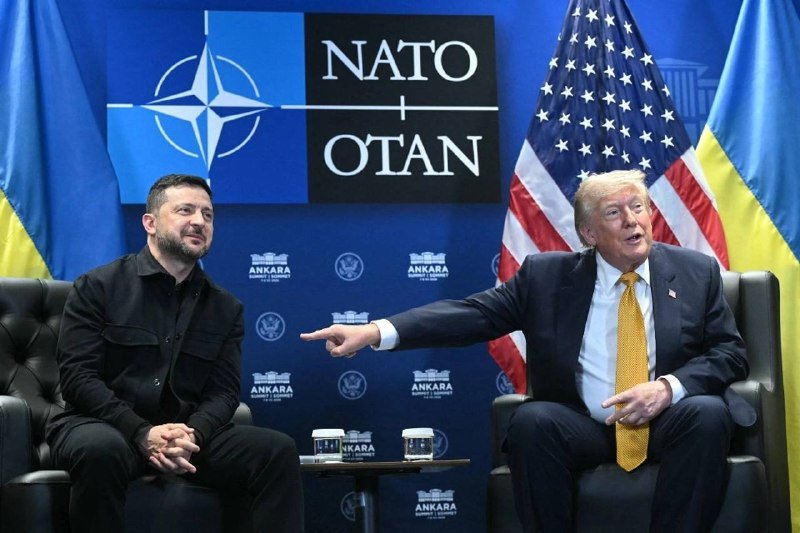 Two men in dark suits sit in leather armchairs at a NATO-branded backdrop with U.S. and Ukrainian flags, one gesturing toward the other.