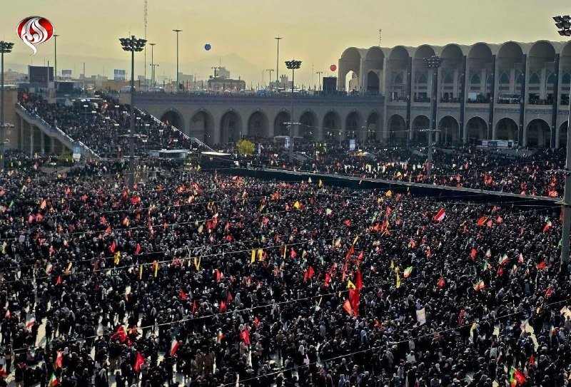 Clerics in white and black robes and turbans stand behind multiple coffins draped in the Iranian flag before a large seated crowd at an outdoor gathering.