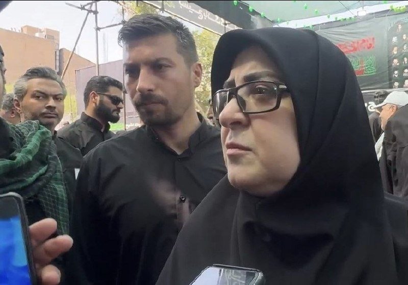 A woman in a black hijab and glasses stands beside a man in black clothing, both looking serious amid a crowd of people at an outdoor gathering.