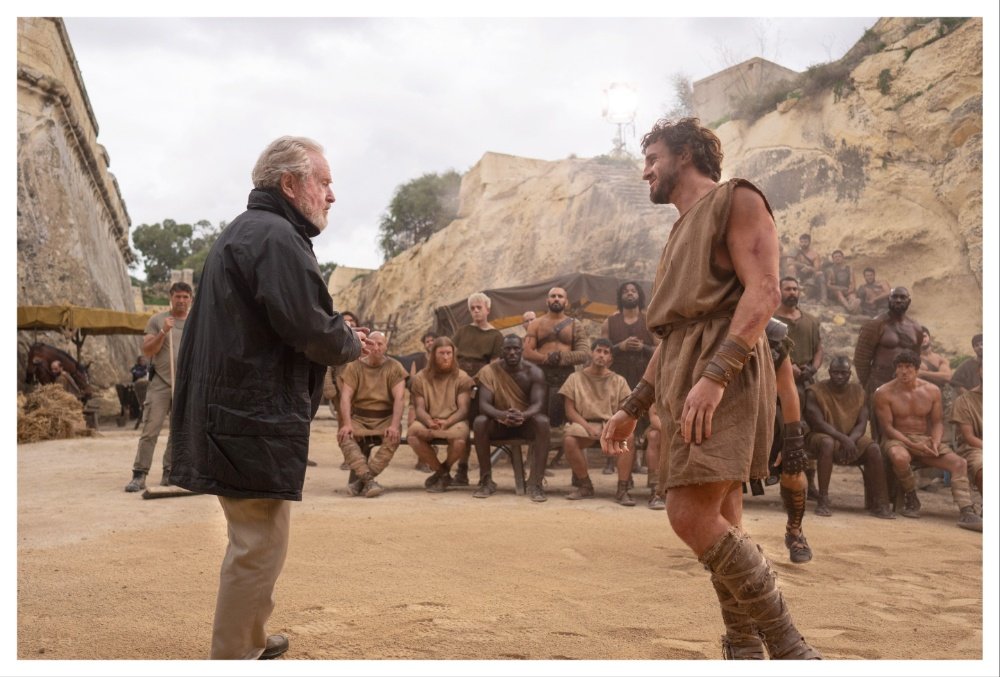 A gray-haired man in a dark jacket converses with a man in a tunic and gladiator sandals on a sandy arena set, surrounded by onlookers.