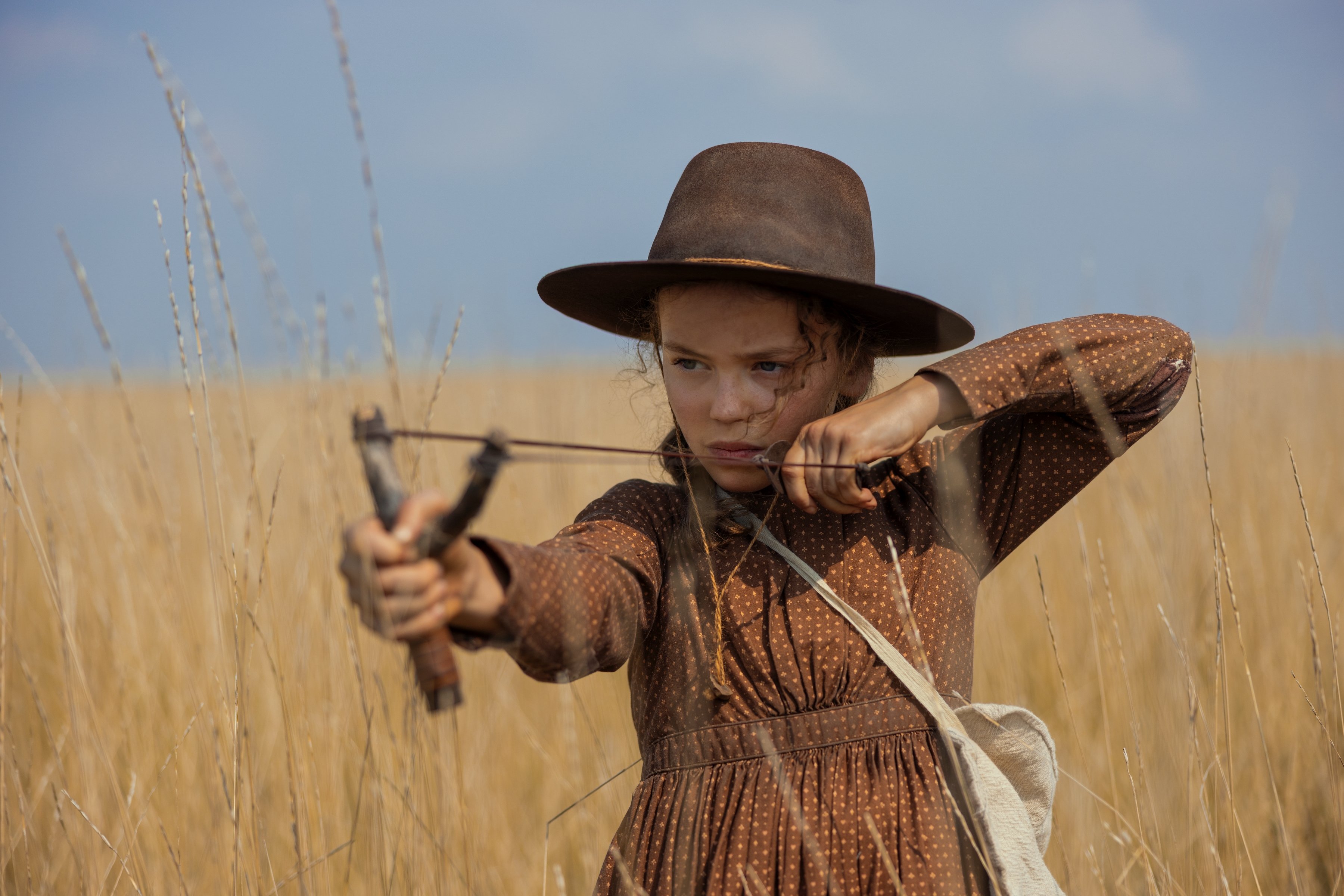 A young girl in a brown hat and dress aims a slingshot while standing in a tall grassy field.