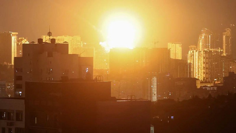 A bright orange explosion illuminates the night sky behind an urban skyline of silhouetted high-rise buildings and rooftops.