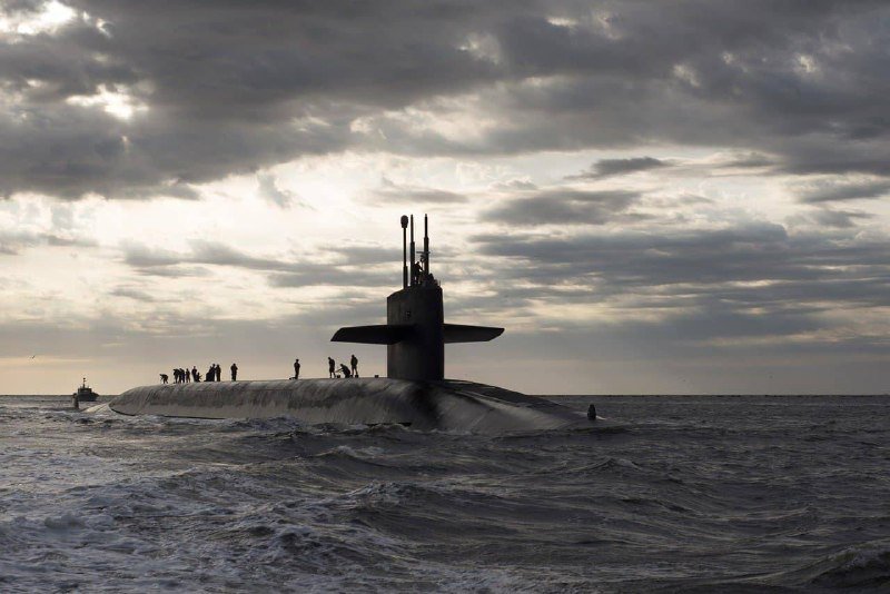 A large submarine floats on choppy ocean waters under a cloudy sky, with several silhouetted figures standing on its deck near the conning tower.