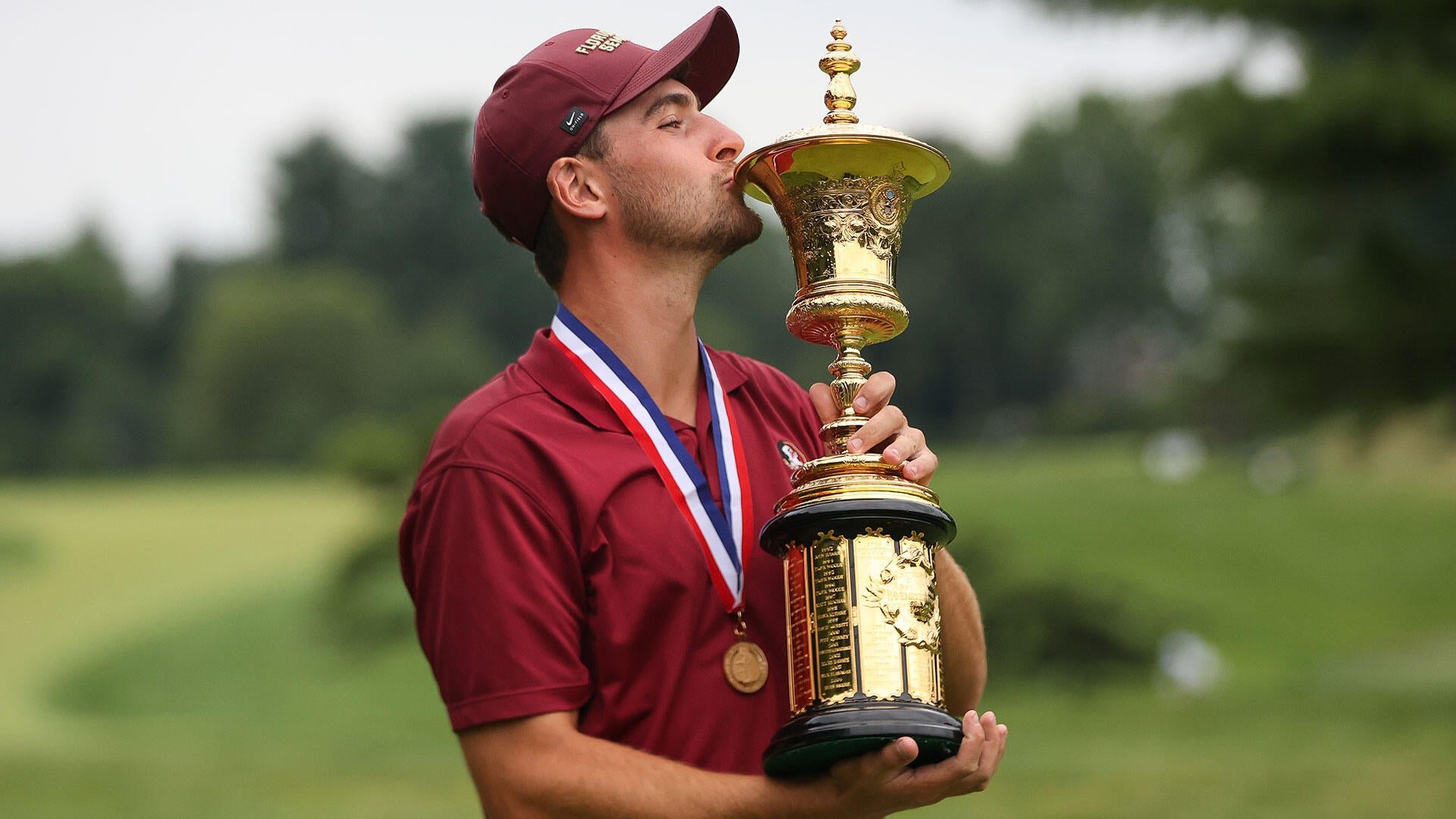 A man in a maroon cap and polo shirt kisses a large gold trophy while wearing a medal, standing outdoors against a grassy field.