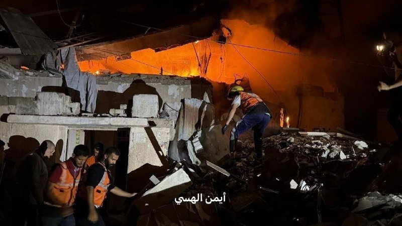 Rescue workers in orange vests sift through the rubble of a collapsed building at night, with flames and thick smoke visible in the background.