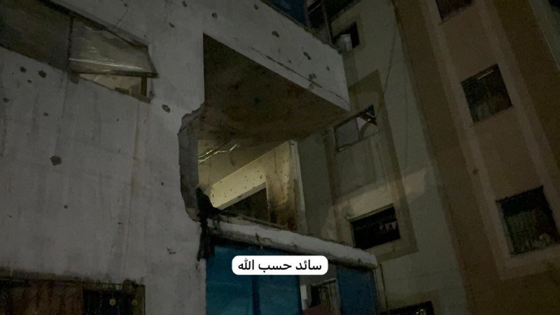 A damaged multi-story building with shattered walls, holes, and exposed concrete is shown at night, featuring Arabic text reading "سأذكِّر حسب الله."