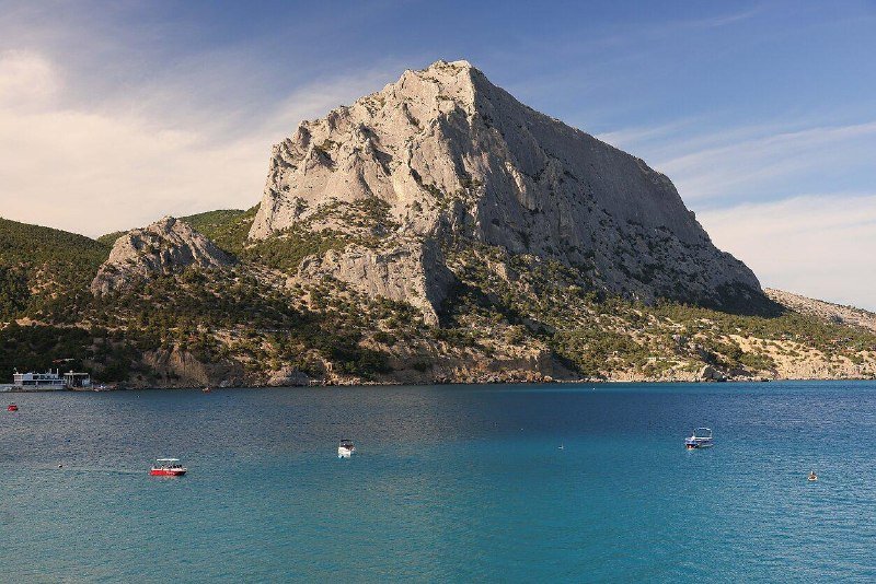 A rocky, pointed mountain rises above a coastal bay where several small boats float on blue water, with a docked structure visible at the shoreline.