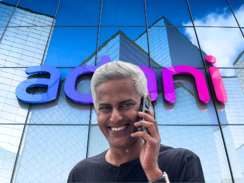 A smiling man with gray hair holds a phone to his ear in front of a glass building displaying a large purple and pink "adani" sign.