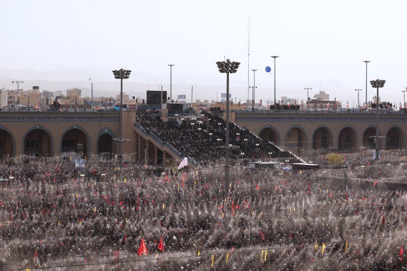 Women in black chadors hold portraits of bearded clerics and wave red flags with Arabic script during a crowded outdoor gathering under a clear blue sky.