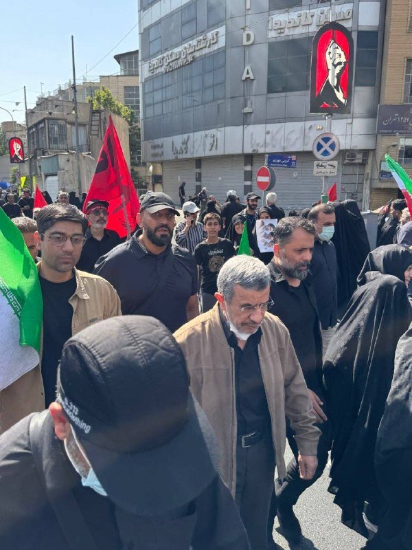 Crowd of people walking down a street carrying flags and holding portraits, with closed storefronts and Persian signage visible in the background.