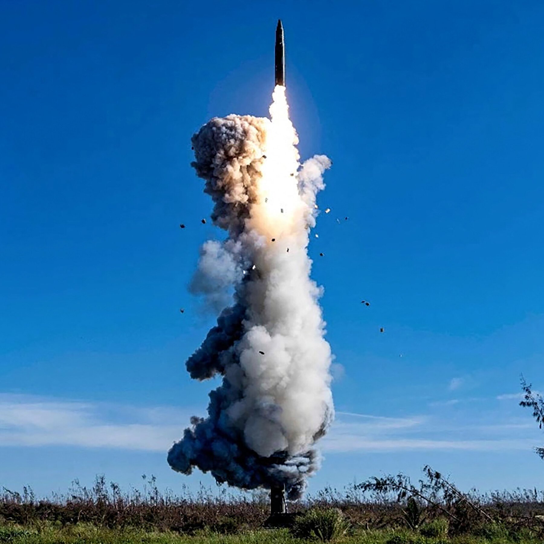 A missile launches from a ground-based launcher, emitting a bright flame and a thick trail of smoke and debris against a clear blue sky above a grassy field.