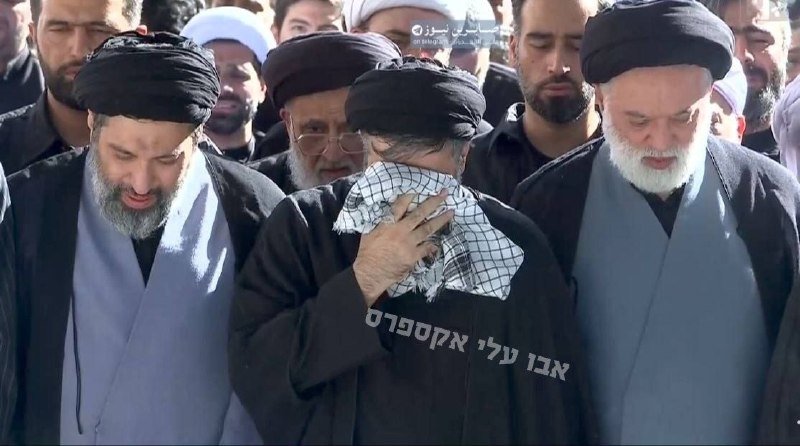 A large crowd of people marches through a city street waving red flags, Iranian flags, and portraits of a turbaned figure, with a Tasnim News watermark visible.