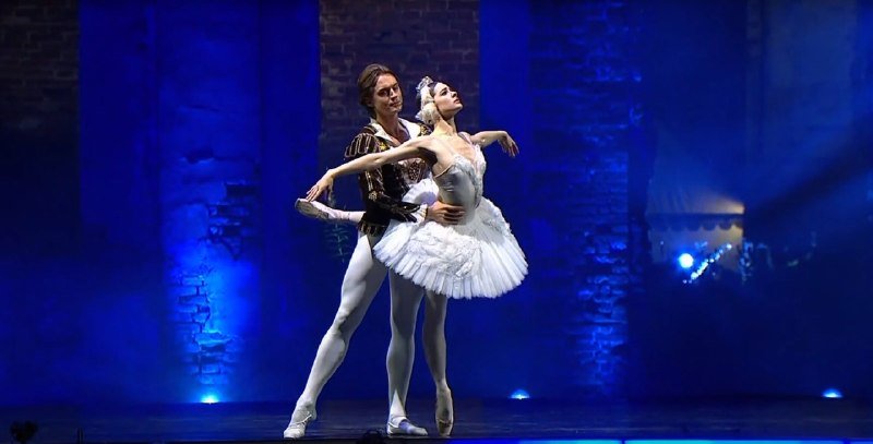 Two ballet dancers perform on a blue-lit stage, the male dancer in a dark tunic and tights supporting the female dancer en pointe in a white tutu.