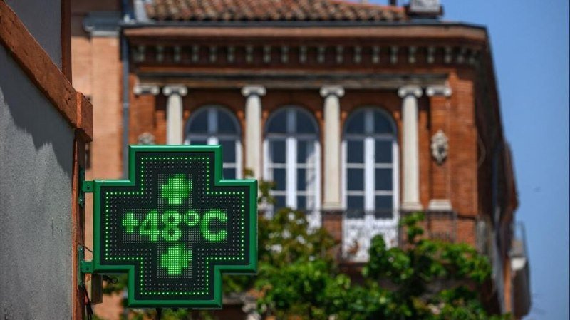 An illuminated green pharmacy cross sign displays "48°C" in the foreground, with a brick building featuring arched windows and classical columns visible behind it.